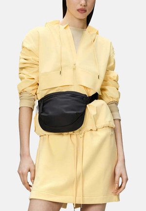Borsa in nylon nera con una superficie liscia, caratterizzata da un design curvo e una tasca frontale con zip, indossata su una tuta leggera gialla.