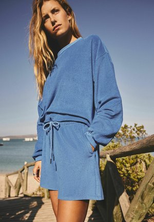 Femeie cu păr lung, purtând un top albastru cu mâneci lungi și pantaloni scurți asortați, stând în aer liber lângă un gard de lemn, aproape de apă.