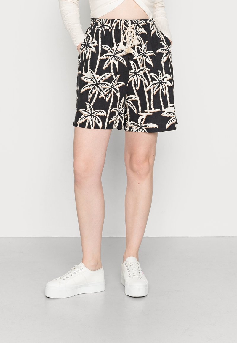 Femme portant un short noir avec un imprimé beige de palmier et des baskets blanches, se tenant devant un fond blanc uni.