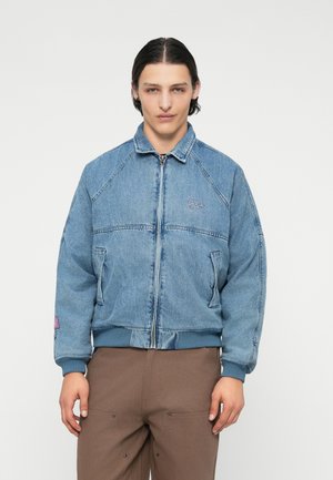 Giacca di jeans azzurro chiaro con chiusura a zip, tasche laterali e polsini a coste. Presenta un logo sul petto e una superficie liscia.