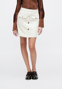 Mini-jupe blanche avec deux poches frontales, fermeture à boutons et texture lisse. Associée à une chemise en satin marron et des mocassins foncés.