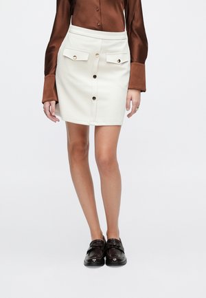 Biała mini spódnica z dwoma przednimi kieszeniami, zapięciem na guziki i gładką fakturą. Do tego brązowa satynowa koszula i ciemne loafersy.