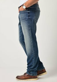 Pantalones vaqueros de mezclilla azul oscuro con parte delantera desgastada, diseño de pierna recta, estilo de cinco bolsillos, combinados con zapatos de cuero marrón.