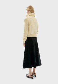 Donna che indossa un maglione beige a collo alto, una gonna midi nera e delle scarpe slingback nere con tacco basso, in piedi di spalle su uno sfondo grigio.
