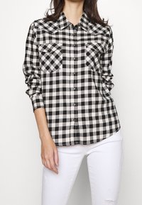 Chemise à carreaux noire et blanche avec deux poches poitrine, fermeture boutonnée sur le devant et manches longues. Fabriquée en tissu doux. Portée avec un jean blanc.