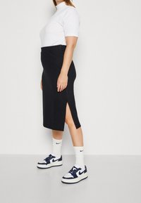 Mustassa ribbimäisessä midi-hameessa on sivuläppä, ja se on yhdistetty valkoiseen tiukkaan poolopaitaan ja laivastonsinisiin lenkkikengiin. Materiaalista johtuva rakenne on näkyvissä designissa.