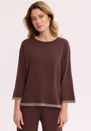 Blondynka z lekkim makijażem nosi luźny brązowy długi rękaw i dopasowane spodnie z paskami na mankietach i na dole.