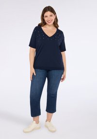 Top navy a maniche corte con scollatura a V e decorazioni colorate sulle spalle, abbinato a pantaloni cropped in denim scuro e sneakers chiare.