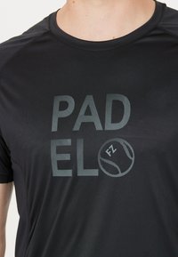 Svart tygsporttröja med "PADEL" och en bolls grafiskt motiv i ljusgrått. Kort ärm, rund halsringning och slät yta.