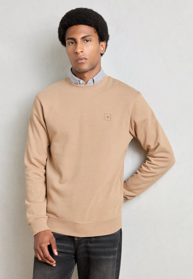 Sweatshirt beige avec des poignets et un ourlet côtelés. Présente une petite étiquette de marque sur la poitrine. Porté par-dessus une chemise à col rayé clair.