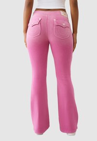 Różowe spodnie dresowe o rozkloszowanym kroju mają gładką fakturę, ozdobne szwy oraz tylne kieszenie zapinane na guziki.