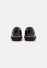 Chaussures en cuir verni noir avec un bout rond, semelle noire plate et un accent beige le long de la semelle. L'arrière présente un petit détail de logo.