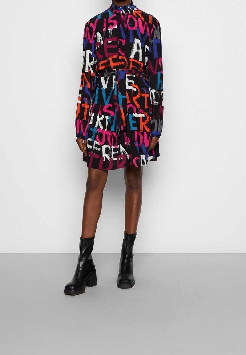 Femme portant une robe multicolore à manches longues avec de grandes lettres en gras et des bottines noires à talons, debout contre un mur blanc.
