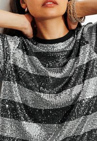 Haut en sequins noirs et argentés avec un col rond. Présente des rayures horizontales et une surface texturée. Accentué par une bordure côtelée noire.