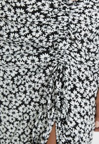 Blumenkleid mit schwarzem Hintergrund und weißen Gänseblümchenmustern, gerafftem Stoff und einem seitlichen Schlitz für zusätzlichen Designakzent.