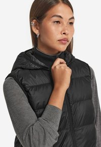 Czarna kamizelka puchowa z kapturem, posiadająca pikowaną teksturę i zamek z przodu. Nosi się ją na ciemnoszarym swetrze z golfem.