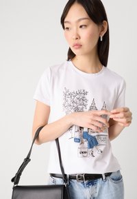 T-shirt en coton blanc avec un design graphique représentant des bâtiments et une silhouette. Associé à un jean bleu clair et un sac bandoulière noir.