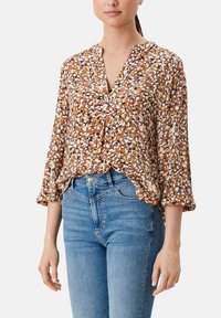 Brun blomsterbluse med V-udskæring, lange ærmer og samlesømme, prydet med et flerfarvet mønster på lette materialer. Bæres med blå jeans.