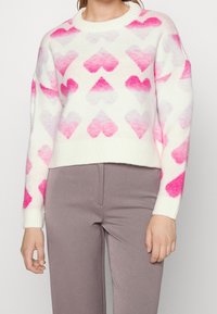 Strickpullover in Creme mit einem Farbverlauf von Rosa in Herzmustern. Verfügt über einen runden Ausschnitt und gerippte Bündchen, die einen lockeren Sitz bieten.