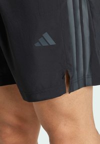 Pantalones cortos deportivos negros de textura suave, con un logotipo triangular gris y tres rayas verticales en el lateral. Aberturas laterales en el dobladillo.