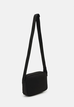 Sort rektangulær crossbody-taske med justerbar rem med subtilt præget tekst, vist mod en ensfarvet lys baggrund.