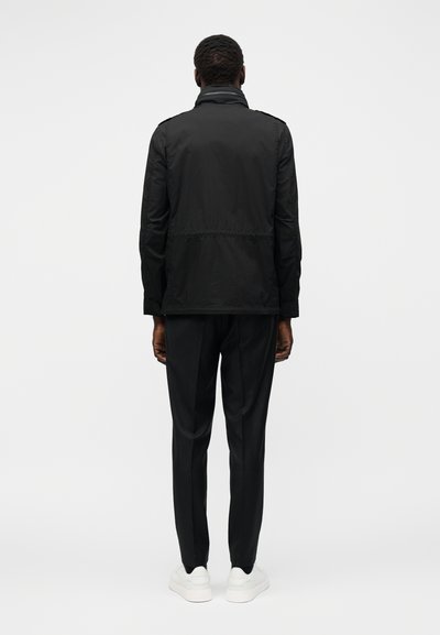 Άνδρας που στέκεται γυρισμένος με την πλάτη φοράει μαύρο μπουφάν, μαύρο παντελόνι και λευκά αθλητικά παπούτσια μπροστά σε απλό φωτεινό φόντο.