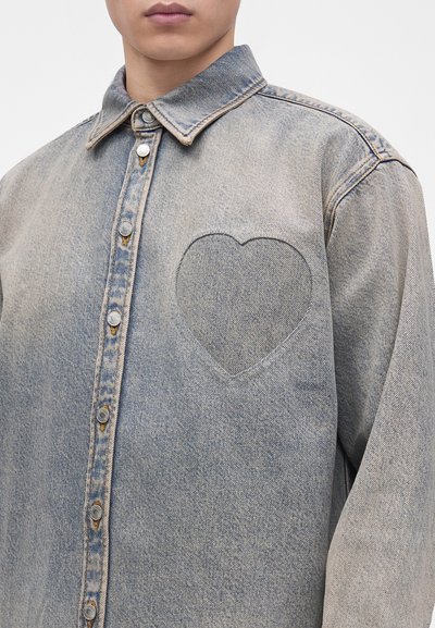 Gros plan d'une personne portant une chemise en denim bleu délavé avec un détail cousu en forme de cœur sur la poitrine et des boutons-pression métalliques à l'avant.