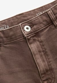 Jeans din denim maro cu o textură netedă, dotate cu un sistem de închidere cu nasture metalic și cusături contrastante. Eticheta este vizibilă pe talie.