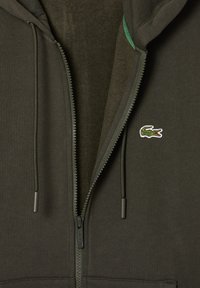 Sweat-shirt zippé à capuche en tissu vert foncé avec une doublure texturée, arborant un petit logo en croix brodé et des cordons de serrage à tige.