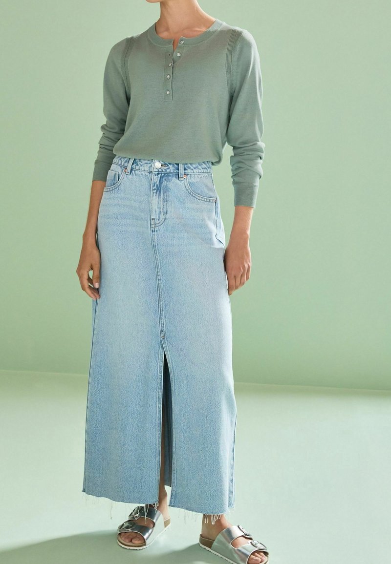 Personne portant un haut henley vert sauge à manches longues, une jupe en jean bleu clair taille haute avec une fente devant et des sandales à boucle argentée.