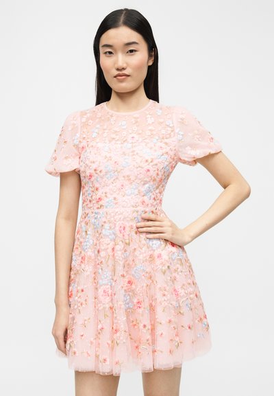 Γυναίκα που φοράει κοντό ρόζ φόρεμα με φουσκωτά μανίκια, διακοσμημένο με πολύχρωμη κεντητή ανθοδέσμη, στέκεται με το ένα χέρι στη μέση.