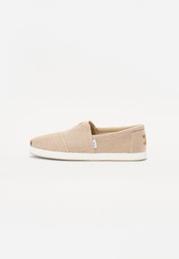 Zapato slip-on de color marrón claro con tejido texturizado, franjas diagonales y suela de goma blanca. Presenta una pequeña etiqueta de logo en el costado.