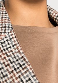 Tkany w kratę blazer w odcieniach brązu, czerni i bieli, w komplecie z gładką beżową bluzką. Posiada wyraźny kołnierz oraz teksturowane wzory.