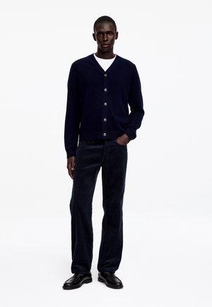 Homme portant un cardigan bleu marine boutonné sur une chemise blanche, pantalon sombre et chaussures noires, debout avec une main dans la poche sur fond blanc.