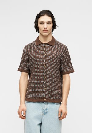 Giovane che indossa una camicia marrone a maniche corte con motivo, bottoni e colletto, e jeans di denim azzurro chiaro, in piedi davanti a uno sfondo semplice.
