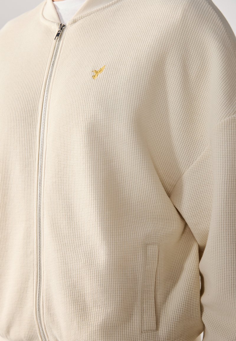 Cremfarbene Jacke mit Waffelstruktur und Reißverschluss, ausgestattet mit einem dezenten goldenen Stickdetail und einem glatten Kragen. Inklusive Seitentaschen.
