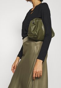 Grön läderhandväska med rynkad design, hålls under armen, i kombination med en svart långärmad topp och en olivgrön satinskjorta.