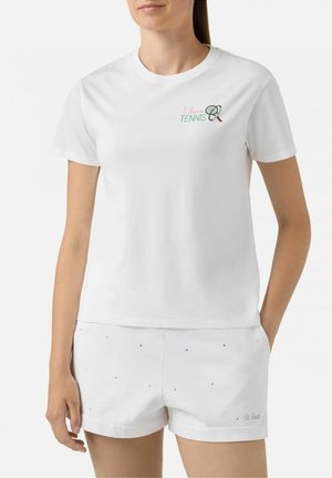 Donna che indossa pantaloncini da tennis bianchi e una maglietta bianca con un piccolo testo "I love tennis" e una grafica di racchetta da tennis sul petto.