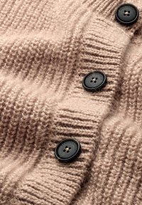 Beige gebreid vest met geribbelde textuur en drie zwarte knopen met de merknaam. Zachte, warme stof.