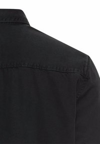 Chemise noire avec un col classique, fabriquée en tissu lisse, présentant une finition soignée et des coutures renforcées aux emmanchures.