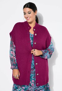 Magenta gestrickte Strickjacke mit kurzen Ärmeln und silbernen Knöpfen, über einem Blumenkleid mit türkisen und rosafarbenen Tönen getragen. Strukturiertes Strickdetail.