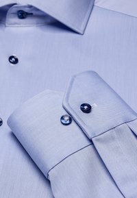 Lichtblauw overhemd van stof met een gladde textuur, met een puntige kraag, contrasterende donkerblauwe knopen en net afgewerkte manchetten.