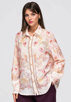 Femme aux cheveux bruns et raides portant une chemise claire à motifs floraux avec des boutons et un pantalon foncé, debout avec une main dans la poche, sur un fond gris.