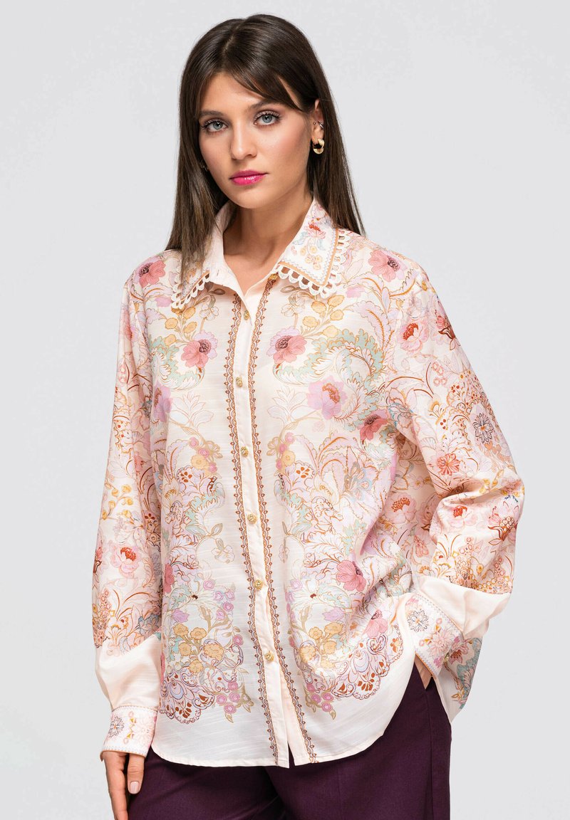 Femme aux cheveux bruns et raides portant une chemise claire à motifs floraux avec des boutons et un pantalon foncé, debout avec une main dans la poche, sur un fond gris.