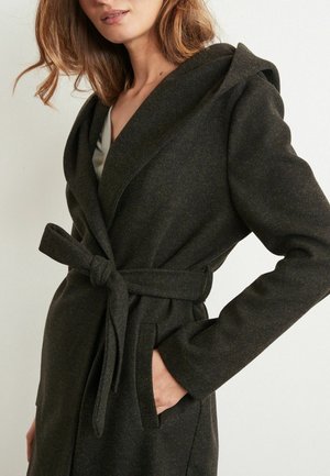 Tmavě zelený vlněný kabát s šálovým límcem, páskem v pase a bočními kapsami. Má měkkou texturu a přizpůsobený střih.