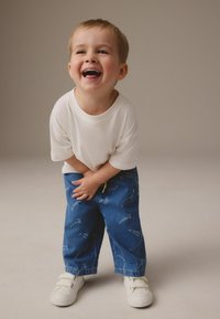 Dziecko ubrane w biały bawełniany t-shirt i niebieskie dżinsowe spodnie z nadrukiem fraz. Buty to białe sneakersy na rzepy.