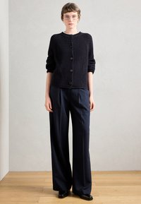 Ναυτικό πλεκτό ζακέτα με κουμπιά, συνδυασμένη με παντελόνι wide-leg ναυτικής ρίγας και μαύρα παπούτσια, σε ουδέτερο φόντο.