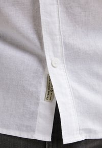 Chemise en lin blanc avec fermeture à boutons, texture subtile et petite étiquette de marque cousue sur la couture latérale.