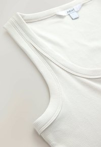 Gros plan sur l'encolure et la bretelle d'un débardeur blanc côtelé avec une étiquette de marque Next visible sur un fond lisse.