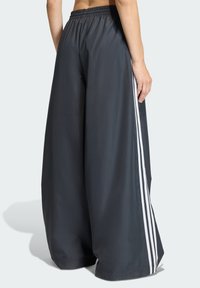 Pantalones deportivos de pierna ancha en gris oscuro, con cintura elástica y tres franjas verticales blancas a lo largo de los lados. Textura de tela suave.